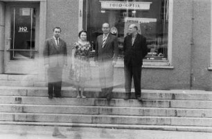 Niesky. Mehrere Personen (Lehrer oder Dozenten ?) auf den Stufen vor einem HO-Industriewarenladen "Foto - Optik"