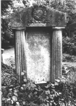 Grabstein 334 (Index: 100082410) Material: Kalkstein Zustand: stark verwittert mit Textverlust Maße HxBxT: 180x200x33 cm Beschreibung: Ädikula mit dorischen Säulen, breiter Sockel, Giebel Symbolik: Kranz mit Schleife, Sterne: Grabstein 334