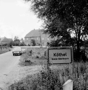 Ortseingang mit Ortsschild: links Pkw: im Hintergrund Bauernhäuser und Bäume