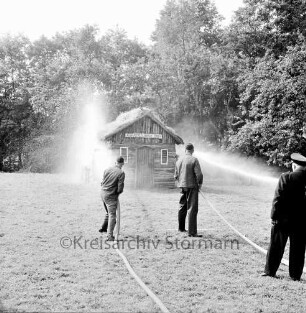 Kupfermühle: Amtsfeuerwehrfest: Löschen der "Kupfermühle Anno 1500": vorn zwei Feuerwehrmännder mit Wasserschläuchen: rechts Wehrführer: hinten brennendes Holzhäuschen: dahinter Bäume