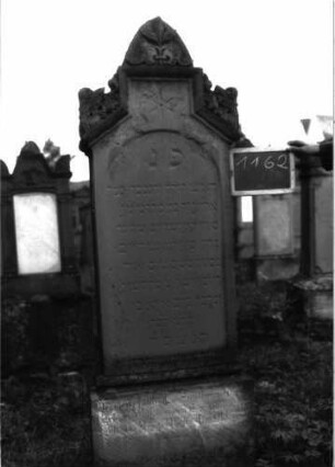 Grabstein 1162 (Index: 100104288) Material: Sandstein Zustand: stellenweise leicht verwittert Maße HxBxT: 190x51x22 cm Beschreibung: Stele auf getrepptem Sockel, giebelartiger, rechteckiger erhöhter Abschluss mit Verdachung, Eck- und Giebelakroterien (Akanthus u. a. Blattornamente), eingetieftes, rundbogiges Schriftfeld, darüber ein eingeritzer Stern Symbolik: Akanthus, Blattornamente, Stern: Grabstein 1162