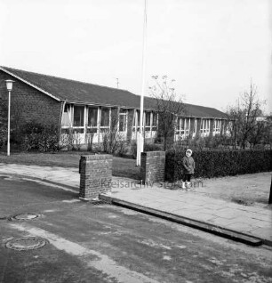 Volksschule: vorn Eingang mit Mauerpfosten und Hecke: rechts davor Kind: dahinter Grünanlage mit Fahnenmast: hinten Pavillonbauten