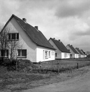 Neubausiedlung nach Fertigstellung: vorn links unbebautes Gelände mit Sträuchern: dahinter Einfamilienhäuser mit Vorgärten