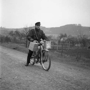 Briefträger mit einem Fahrrad auf einer Landstraße, am Lenker hängen zwei Pakete