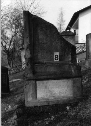 Grabstein 8 (Index: 100080871) Material: Sandstein mit weißer Marmorplatte im Sockel Zustand: Inschriftenplatte der Stele fehlt, rechter Teil abgebrochen Maße HxBxT: 98x57x24 cm Beschreibung: ädikulaartige Stele, getreppter Sockel: Grabstein 8