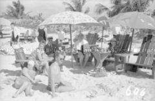 Touristen in Badekleidung am Strand unter einem Sonnenschirm sitzend