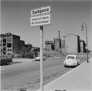 Hinweisschild "Sackgasse verursacht durch die Schandmauer". Im Hintergrund Grenzsicherungsanlagen der DDR