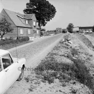 Straße mit Kopfsteinpflaster: links Wohnhaus mit Holzstapel: rechts planiertes Gelände mit Markierungspfählen: hinten Mitte Siedlungshäuser, rechts Bauernhof: vorn links Pkw