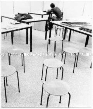 Hinten rechts sitzt ein Mann an einem Tisch, neben ihm weitere Tische, vorne stehen sechs runde Hocker