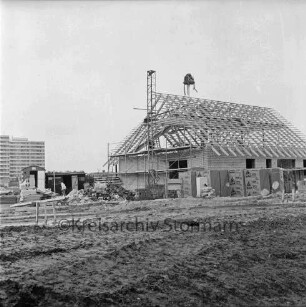 Dammannsche Koppel: Neubaugebiet: vorn unbefestigtes Gelände: rechts Einfamilienhaus mit Gerüst und Richtkranz: davor Baumaterial: links Bauwagen und Arbeiter: hinten links Hochhäuser am Poggenseer Weg: 15. Oktober 1976