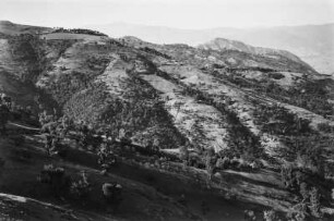 Dorf und Landschaftsblick : Blick auf das Dorf At Hicem, das auf einer Bergkuppe gelegen ist. An den Berghängen lassen sich Gerste- und Weizenfelder sowie Feigenbäume, Macchiebüsche, Eichen und Eschen erkennen.
