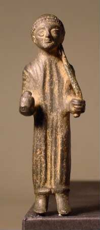 Rohe archaische Statuette einer stehenden, langbekleideten Figur; unbärtig; um den Kopf eine doppelte Flechte; rechts ein Granatapfel; links ein Stab