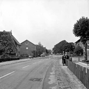 Alte Dorfstraße: vorn rechts Zaun: daran zwei Frauen: hinten Bauernhäuser und Grünanlage
