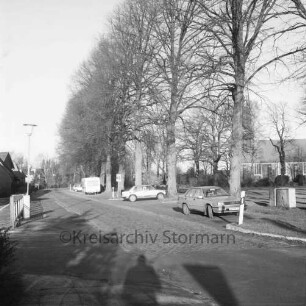 Kirchenweg: Kopfsteinpflaster: in der Mitte in Parkbuchten Pkws und Wohnwagen: dahinter Baumreihe des Dorfangers: hinten rechts Friedenskirche und Kirchhof: links Straßenlampe und Häuser