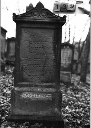 Grabstein 139 (Index: 99751950) Material: Sandstein Zustand: abgeblättert mit großem Textverlust Maße HxBxT: 148x52x18 cm Beschreibung: St. , Sock. , Abdeckplatte m. Volutenaufsatz m. Palmette, rechteck. Schriftfeld eingetie Symbolik: Voluten, Palmette: Grabstein 139