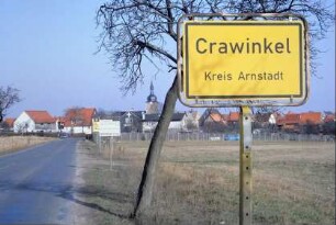 Ortseingansschild; Straße, Baum; dahinter Häuser und Gärten am Ortsrand