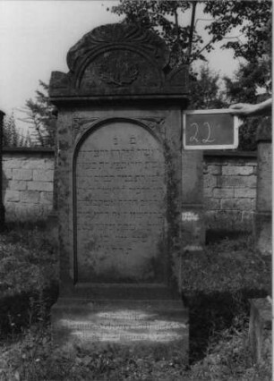 Grabstein 22 (Index: 99801897) Material: Sandstein Zustand: verwittert Maße HxBxT: 153x55x22 cm Beschreibung: Stele, Sockel, Rundbogengiebel mit Eckakroteren, eingetieftes Schriftfeld mit Rundbogen Symbolik: Eichblätter, Blätterornamentik auf dem Rundbogen: Grabstein 22