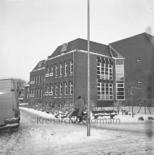 Woldenhorn-Schule: Sonderschule des Kreises Stormarn für geistig behinderte Kinder: Ecke Reeshoop / Schulstraße: Außenaufnahme: im Winter: vorne rechts Einmündung Hans-Schadendorff-Stieg, mit Straßenschild: Maschendrahtzaun, Verkehrsschild, Ruhebank: Person auf Fahrrad, Kleintransporter, Fahnenmast