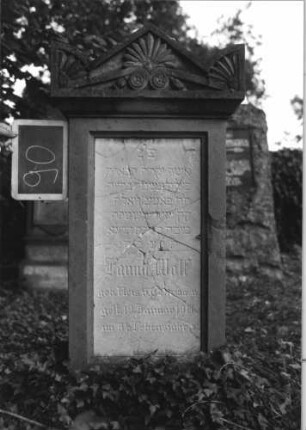Grabstein 90 (Index: 100080556) Material: Sandstein, weiße Marmorplatte Zustand: Platte mehrfach gesprungen Maße HxBxT: 142x50x26 cm Beschreibung: Stele, Giebel m. stilisiert. Eckakroteren, rechteckige Schriftplatte Symbolik: Mohnkapseln, Blätter, Voluten im Giebel: Grabstein 90