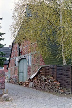 Schulstraße (10): Bauernhaus aus Fachwerk: Giebelseite Wirtschaftstrakt mit Scheunentor, darüber Luke: Stapel mit Holzscheiten: Toreinfahrt: rechts Sichtschutz