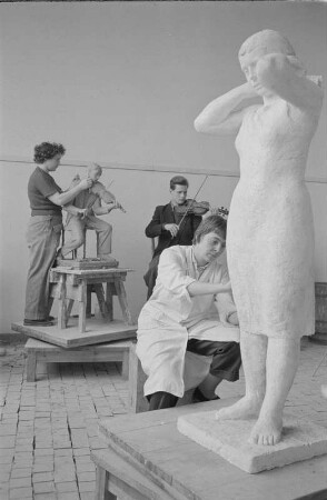 Studenten der Bildhauerklasse von Walter Arnold beim Modellieren. Im Vordergrund die Gips-Statue einerstehenden Frau, ein Kopftuch bindend (1960/1962)