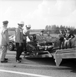 B 208: Kurve bei Westerau: Verkehrsunfall: Zusammenstoß von zwei Pkws: Feuerwehrmänner beim Aufladen von Unfallwagen mit eingedrückter Front auf Plattform von Abschleppwagen: rechts Zuschauer: Zaun, hinten Feld, im Hintergrund Wald