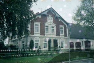 Dorfstraße 10: Bauernhaus: giebelständiges Wohnhaus: Straßenfront mit Eingangstür, Gesims, hölzerne Giebelzier: links Traufseite: rechts Querbau Wirtschaftstrakt: Traufseite mit Toren: im Vordergrund Gartenzaun aus Schmiedeeisen