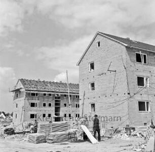 Klosterbergen: Bogenstraße: Wohnblocks im Rohbau, links Dachdeckerarbeiten: vorn Arbeiter zwischen Stapeln von Holzpaletten: rechts und links Ziegelstapel und Baumaterial: hinten links Einfamilienhaus