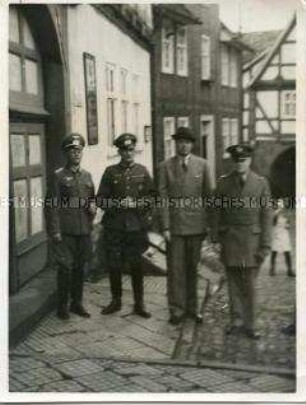 Zwei Offiziere der Wehrmacht, ein weiterer Soldat und ein Mann im Anzug vor Fachwerkgebäuden