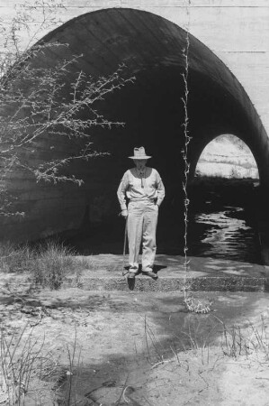 Mann vor einer Brücke : Ein Mann steht vor einer Unterführung, durch die ein Bach fließt.