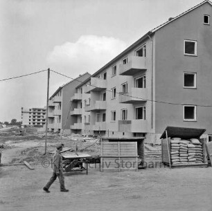 Klosterbergen: Bogenstraße: rechts dreigeschossiger Wohnblock vor Fertigstellung: davor Werkbank und zwei Holzcontainer mit Zementsäcken: vorn links Arbeiter: dahinter Reste von Baumaterial auf unbefestigtem Gelände: hinten links achtgeschossiges Punkthaus im Bau
