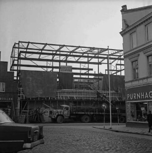 Firma Carl Hoffmann: Hindenburgstraße: Elektrohandel: Neubau: Aufbau des Stahlgerüsts: davor Bauzaun und Lkw: links Wohn- und Geschäftshaus des Haushaltswarengeschäfts Otto Möller: rechts Hausecke des Wohn- und Geschäftshauses mit Drogerie Purnhagen: im Vordergrund Marktplatz, Pkw-Heck