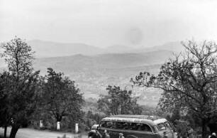 Talblick : Blick in ein Tal, in dem eine Ortschaft liegt. Im Vordergrund ein Bus auf einer Straße, davor drei Personen.