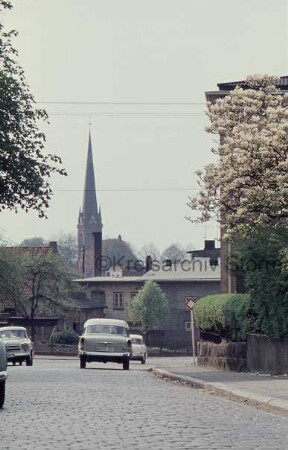 Ratzeburger Straße mit Kopfsteinpflaster: links und Mitte Pkws: rechts Mauer mit Hecke und blühendem Magnolienbaum: hinten rechts Einmündung der Bahnhofstraße: dahinter Gebäude: hinten links Turm der Peter-Paul Kirche