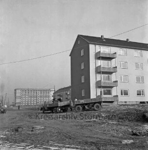 Klosterbergen: viergeschossige Wohnblocks vor Fertigstellung: davor Lkw mit Anhänger: darauf Arbeiter beim Entladen von Baumaterial: hinten links achtgeschossiger Punkthaus mit Gerüst