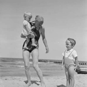 Frau in Bademode mit Kleinkind im Arm und weiteres Kind am Strand