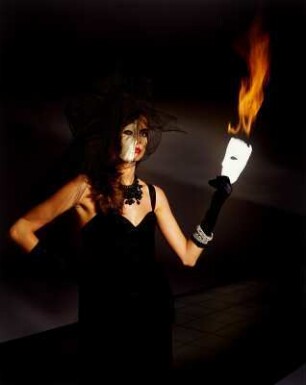 Model in schwarzem Kleid mit weißer Halbmaske und brennender Halbmaske in der Hand