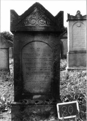 Grabstein 80 (Index: 99936875) Material: Sandstein Zustand: gut Maße HxBxT: 136x53x21 cm Beschreibung: Stele, profilierter Sockel, Giebel mit stilisierten Eckakroteren, Schriftfeld eingetieft mit Segmentbogenabschluß Symbolik: Blüte und stilisierte Ranken im Giebel: Grabstein 80