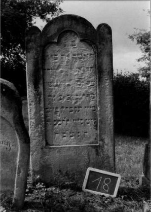 Grabstein 18 (Index: 100107116) Material: Sandstein Zustand: stark verwittert mit Textverlust Maße HxBxT: 131x61x16 cm Beschreibung: Stele, eingezogener Rundbogenabschluß mit stilisierten Eckakroteren, Schriftfeld mit Kleeblattabschluß: Grabstein 18