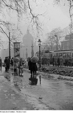 Menschen mit Regenschirmen warten an der Straßenbahnhaltestelle auf dem Martin-Luther-Ring, im Hintergrund das Reichsgericht