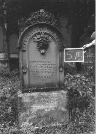 Grabstein 575 (Index: 99895388) Material: Sandstein Zustand: gut, Sockel leicht verwittert Maße HxBxT: 148x59x18 cm Beschreibung: Stele mit Rundbogenabschluss, kleiner Verdachung und Mittelakroter; Sockel Symbolik: Rosengebinde: Grabstein 575 (1)