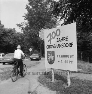 Sieker Landstraße: vorn Hinweisschild auf 700-jähriges Jubiläum der Gemeinde: links dahinter Verkehrsschild: vorn links Fahrradfahrer auf Fahrradweg: dahinter Pkws: hinten Bäume