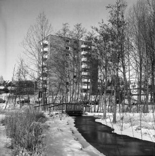 Feuchtgebiet zwischen Masurenweg und Ratzeburger Straße: rechts Bach: in der Mitte Holzbrücke für Fußgänger: dahinter Baumreihe: hinten Garagentrakt mit Pkws: im Hintergrund mehrgeschossiger Wohnblock und Reihenhäuser: Winter