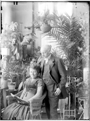 Wohnung der Familie Knochenhauer, Wintergarten? Dame, mit einem Buch im Korbstuhl sitzend, dahinter Herr (Ehemann?) stehend : Kattowitz, Friedensstraße 2. Wohnung der Familie Knochenhauer, Wintergarten? Dame, mit einem Buch im Korbstuhl sitzend, dahinter Herr (Ehemann?) stehend
