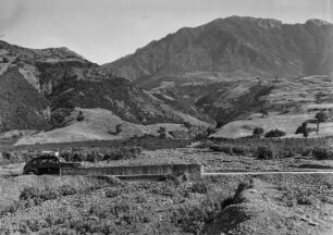 Landschaftsblick : Etwa 25 km östlich an der Küste Bejaias. An den Berghängen befinden sich Macchiebüsche. Im Vordergrund ist eine mit Geröll bedeckte Brücke zu erkennen.