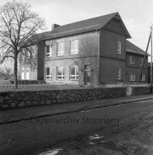 Volksschule mit Erweiterungsbau: links Schulhof mit Baum: davor Mauer mit Zaun: im Vordergrund Dorfstraße
