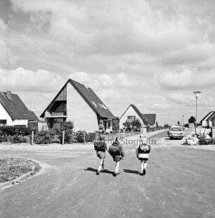 Neubausiedlung: links Einfamilienhäuser mit Vorgärten: rechts hinten Sandhaufen und Pkws: im Vordergrund auf Straße drei Schulkinder