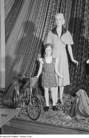Textilien und Damen- sowie Kinderbekleidung an Schaufensterpuppen, dekoriert mit einem Dreirad auf der Leipziger Herbstmesse 1954