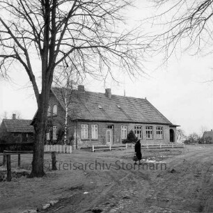 Volksschule: davor Zaun: links Baum: hinten rechts Frau: vorn rechts Straße mit Kopfsteinpflaster