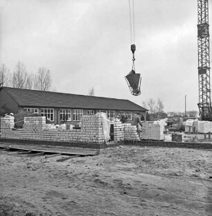 Volksschule: Erweiterungsbau: Bauarbeiter beim Setzen der Grundmauern: rechts Stapel mit Ziegeln: dahinter Baukran, an Ausleger Baumaschine: links Erd- und Sandhaufen: hinten Altbau